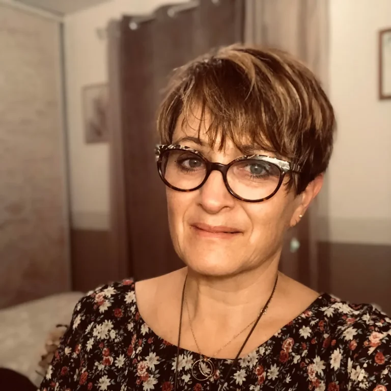 Femme avec des lunettes, cheveux courts et châtain, souriant dans une pièce lumineuse.